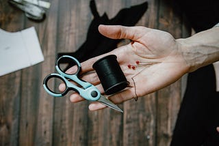 An older person’s wrinkled hand holding out scissors, thread, and pins in the palm of their hand over a work table with fabric scraps and patterns