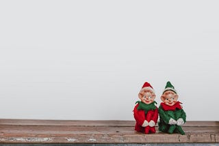 Two toy Santa elves sitting on a table.