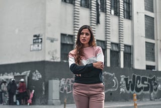 Person with their arms crossed and an annoyed expression standing on a street in front of a wall with graffiti.