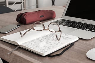 Items are scattered across a wooden desk. Items include a laptop, open notebook, a pair of glasses, and a mobile device.