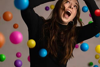 A woman seated looking up hands in the air trying to avoid colorful balls falling on her