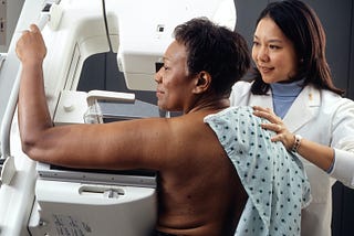 Black women undergoes a mammogram with an Asian women in a white lab coat as the technician.