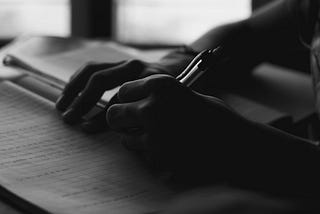 A black and white photo of a person’s hands holding a pen and writing in a notebook.
