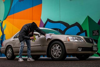 Un hombre limpia su coche con un trapo. El coche es de color marrón y es un modelo de en torno al año 2000. Hay un grafiti colorido tras el coche en el que destacan los colores naranja, azul, verde, y turquesa.