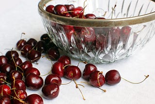 Vintage, crystal bowl with leaded rim, half-full of casually tossed cherries against a white background with more fresh cherries scattered on the white tabletop.