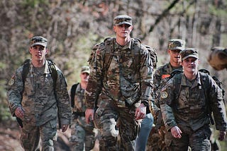 A small group of people in camouflage military uniforms and caps hike down a wooded trail, wearing large backpacks and gear, with relaxed, focused expressions in soft natural light.