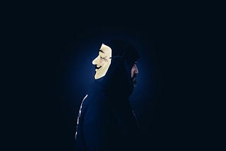 banner image of a man in a hoodie with a mask on the back of the face. Dark blue background. Depicting a developer suffering from developer imposter syndrome.