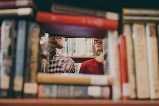 There is a stack of books with a open square in the middle, through it two smiling faces are seen from the nose down. They are facing each other.
