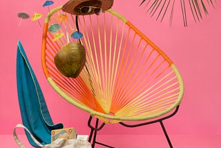 In front of a pink backdrop, an invisible figure, visible only by a hat, shoes and coconut cocktail, reclines on a chair, under a palm tree.