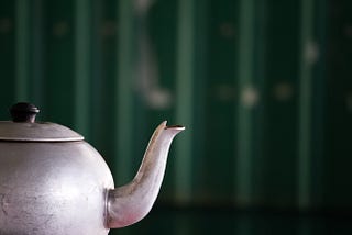 A light silver/grey tea pot is in front of a green background.