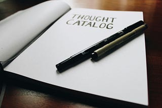 Notebook with the words “Thought Catalog” on them, with a pen and a blank page