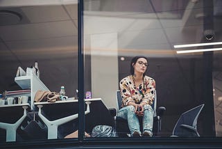 A woman wearing glasses and a floral print top sits alone in a modern office meeting room, viewing outside through a glass partition. She is wearing ripped jeans and sitting casually in an office chair. The room has fluorescent lighting and modern office furniture, with some personal items visible on a desk. The image has a contemplative or pensive mood.
