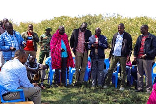 A group of men standing in a group outside with trees in the background. One man wears a traditional red Maasai shuka.