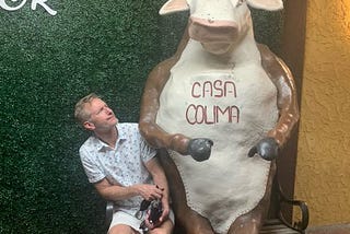 Photo of Ted Wheeler sitting next to plaster cow with Casa Colima written in red on its chest. He is making a silly face.