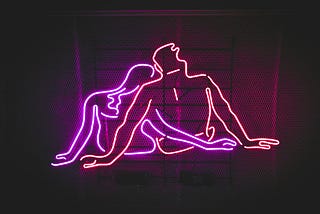Neon sign depicting two abstract human figures in intimate embrace, created with pink and red glowing tubes against a dark mesh background.