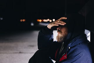 Photo of a man sitting on the ground outside at night with his head in his hand.