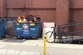 On a city sidewalk is a blue dumpster, bright orange flames peeking out.