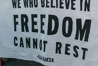 A group of masked people at a protest holding a large banner that reads “We who believe in freedom cannot rest.”