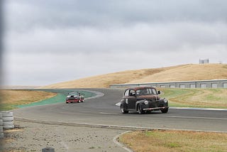 The 1941 Ford Coupe coming out of a curve on the racetrack, followed by a pickup truck.