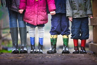 A close-up of four children standing side by side wearing colorful rain boots covered in mud. The lower half of their bodies is visible, showing different pants, leggings, and coats.