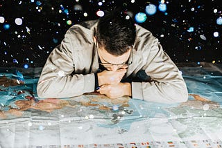 Image of a man with dark hair, glasses and a tan shirt looking down at a desk sized paper map of the world thinking. The image was chosen by Spiritual Lifestyle Writer The Christine McDonald to depict a person searching for their life purpose
