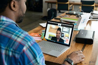 Man in button-up shirt with laptop open on the table to a female interviewer on the screen.