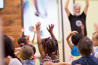 This image features a group of young children in a classroom, with several of them raising their hands high. In the background, an adult with gray hair is also raising his hands, suggesting he might be leading the group in an activity. The focus is on the children, who are engaged and appear to be having fun.