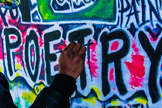 a man’s hand using a brush to finish painting the word ‘poetry’ in a colourful graffiti style