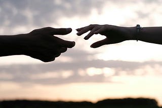 Silhouettes of two hands reaching toward each other against a cloudy evening sky, moments before their fingertips meet.