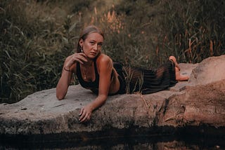 An attractive girl lies sprawled on a rock by some water, woodland in the background.