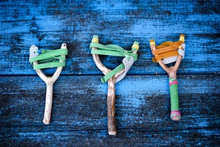 Three slingshots made from wood and rubber bands are laid out in a row on a blue painted wood table.
