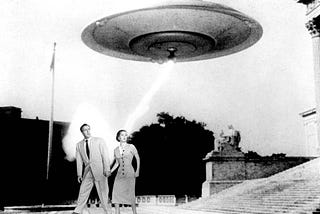 A black and white photo of a saucer-shaped UFO hovering above a white man and white woman in 1950s style clothing.