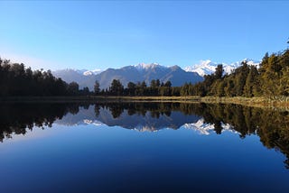 A sunlit mountain sits behind a lake, which is surrounded by trees