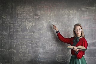 A lady in the red dress is standing in front of a blackboard pointing towards an equation on the board while facing the camera