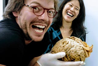 The author holds a brain in the foreground while laughing fiendishly, as Dr. Suzuki laughs from behind.