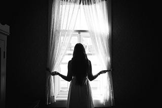 A rear view of a woman looking out through a curtained window
