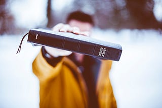 A man in a yellow coat holding a Holy Bible out toward the camera