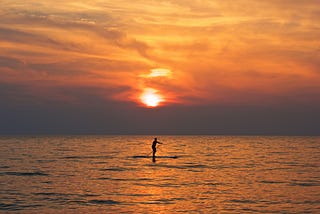 A person paddleboarding on the sea during sunset with a golden sky and a the sun that nearly set in the background.