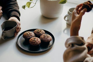 two people sitting around a table with coffee and muffins