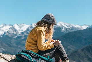Female hiker sits on a mountainside with her journal in her lap and pack by her side.