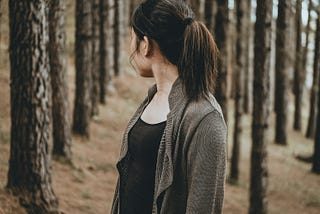 A brunette with a ponytail and an oversized cardigan is in the forest looking back at the trees.