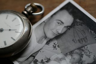 The image shows a vintage pocket watch with Roman numerals on its face lying next to an old black-and-white photograph. The photograph features a smiling young man and a baby, possibly suggesting a family connection or a memory from the past. The items are placed on a wooden surface, giving the scene a nostalgic and timeless feel.