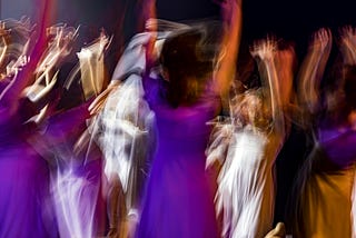 A photo of people dancing with arms in the air in a dark room. There is a lot of motion depicted due to an extended exposure and some figures are brightly lit (white), others less so (purple).