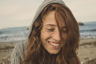 An auburn haired, freckled woman wearing a grey knitted hoodie. She’s on a beach, smiling at something down and away from the camera lens.
