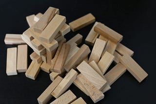 A heap of wood jenga blocks. Most have been thrown onto a black surface; a few are haphazardly stacked on top of each other.