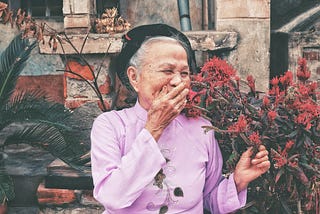 An elderly woman in a purple outfit, smiling while next to a bush of red flowers.