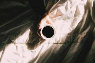 Picture of a mug of coffee in bed with morning sunrise shining through.