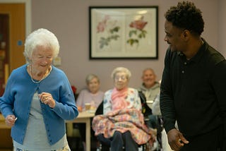 A senior in a senior-center environment dances next to a young adult