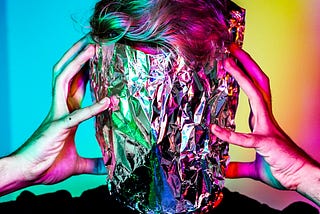 A technicolor image of a masculine-presenting person with short blonde hair holding his outstretched hands to the side of his face. His face is covered in tin foil, which reflects the blue and yellow wallpaper to look green and pink. Only the upper shoulders are visible, and the man is wearing a black tee.
