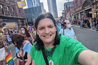 Chris Northwood, smiling, with a trans flag on her cheek, walking down Deansgate with towers in the background and crowds onlooking with a group of others around her